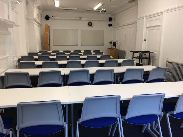 Flat floored teaching room with rows of tables and chairs, whiteboards, screen, and projector