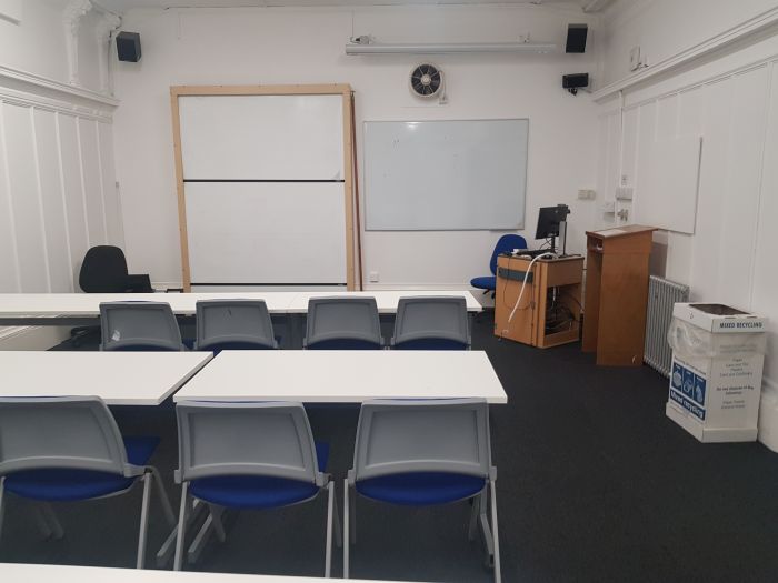 Flat floored teaching room with rows of tables and chairs, whiteboards, screen, and projector