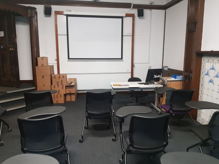 Flat floored teaching room with rows of tablet chairs, whiteboard, screen, lecturer's desk and chair, and PC.