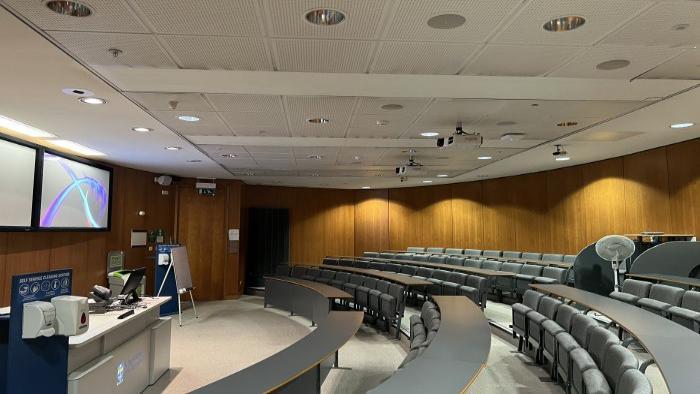 Raked lecture theatre with fixed seating, projectors, large screens, PC, and lectern.