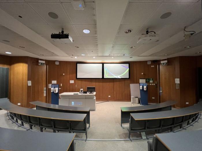 Raked lecture theatre with fixed seating, projectors, large screens, PC, and lectern.