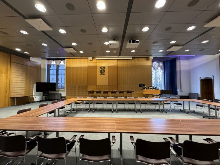 Flat floored room with tables and chairs in a boardroom layout, raised podium with tables and chairs in a row, projector, lectern, video monitor, and PC.