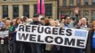 photograph of a mixed ethnic group of people of different genders, holding up a white banner with black text that reads wrefugees welcome. the background seems to be city centre of Glasgow 
