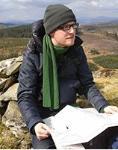 photo of Ben Colburn: a young white man with blonde facial hair and square glasses, wearing a dark blue puffer jacket, green scarf and green hat kneels in a rocky landscape with a map on his lap