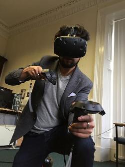 photo of a man in jeans, a blazer and white tshirt, with black beard and curly hear, kitted out with a virtual reality headset and two handheld controllers, moving towards the camera. in the background there are wires and a computer set up in a classroom