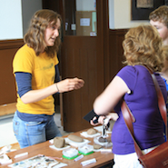 A woman speaking to two people. Archaeological finds are on the table in-between them.