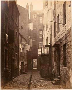 A sepia toned photograph of a narrow cobble stoned street between two blocks of tenement buildings.  Washing hangs out of some of the windows.  The impression is of poverty and poor living conditions