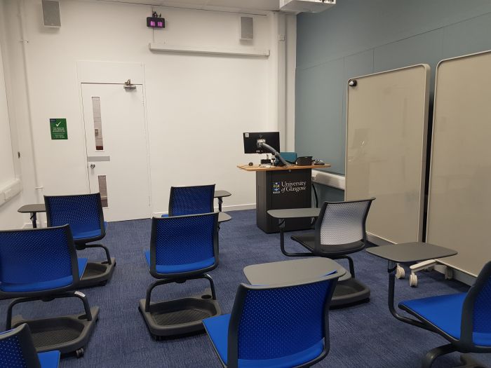 Flat floored teaching room with tablet chairs, projector, large screen, PC, lectern, and movable glassboards.