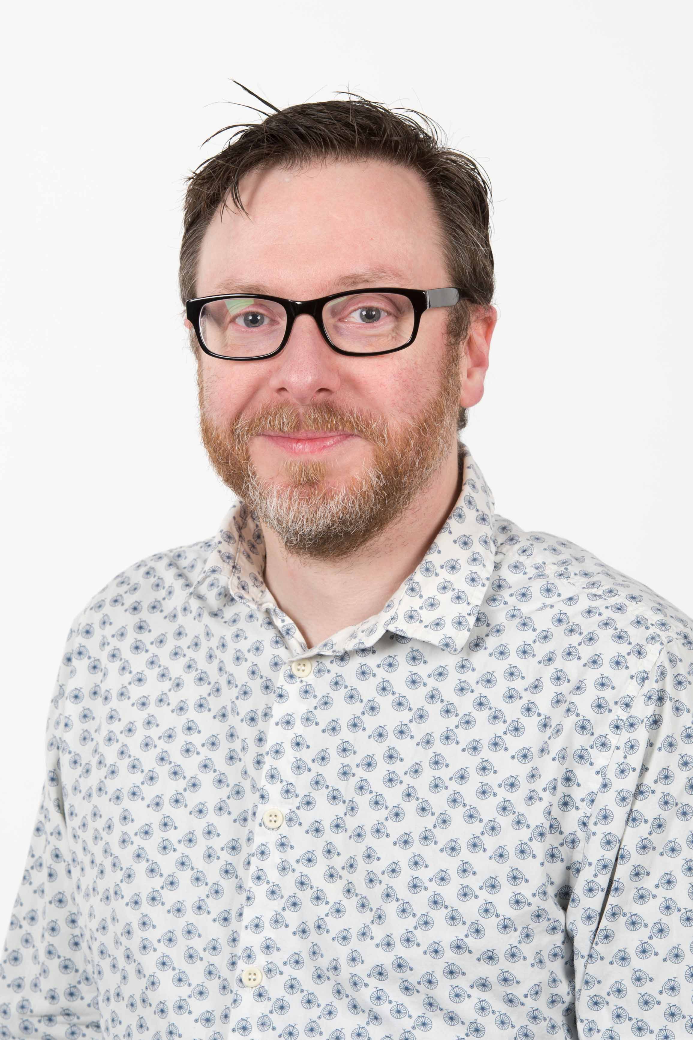 photo of Chris Lindsay, a middle-aged white man with short brwon hair and short ginger beard, wearing dark-rimmed square glasses and a white shirt with small print of unicycles