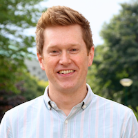 photo of keith wilson, a young white man with light brown short hair sweeped back, wearing a light blue striped dress shirt, with greenery in the background