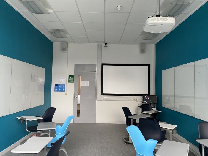 Flat floored teaching room with tablet chairs, PC, projector, large screen, and whiteboards.