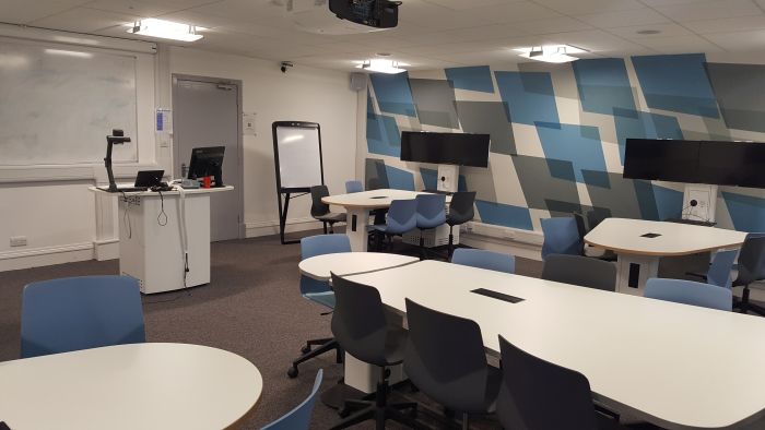 Flat floored Tech Enabled Active Learning (TEAL) room with groups of tables and chairs, each with a video monitor, as well as whiteboard, projector, moveable whiteboard, visualiser, PC, and lectern.