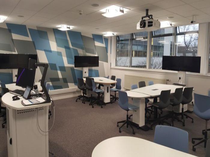 Flat floored Tech Enabled Active Learning (TEAL) room with groups of tables and chairs, each with a video monitor, as well as PC, lectern, visualiser, and projector.