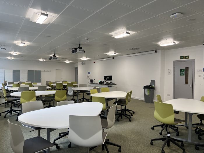 Flat floored teaching room with groups of teardrop tables and chairs, projectors, PC, and lectern.