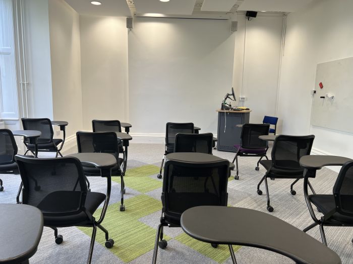 Flat floored teaching room with tables and chairs in horseshoe set-up, screen, and PC