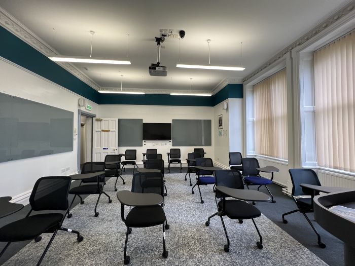 Flat floored teaching room with tables and chairs in round table set-up, glassboards, and video monitor