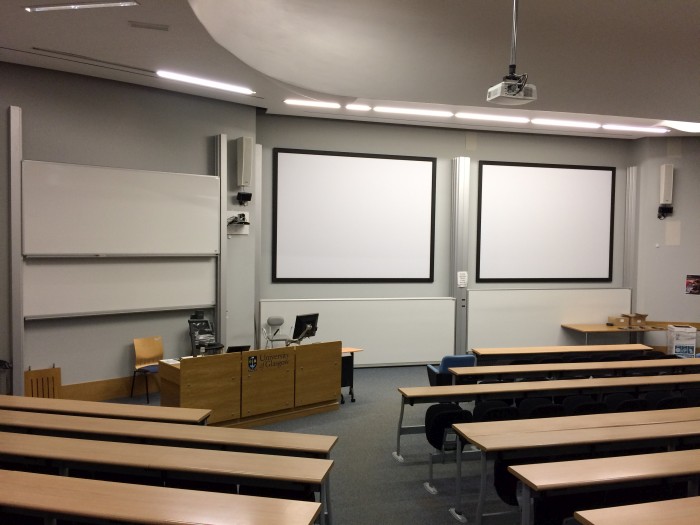 Raked lecture theatre with fixed seating, whiteboard, screens, PC, and projector