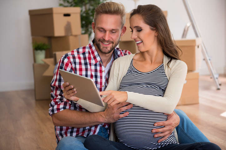Young pregnant couple viewing tablet device, with boxes behind them, 