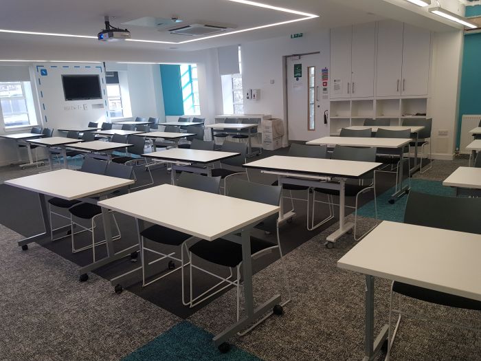 Flat floored teaching room with rows of tables and chairs, writing walls, video monitor, and projector.