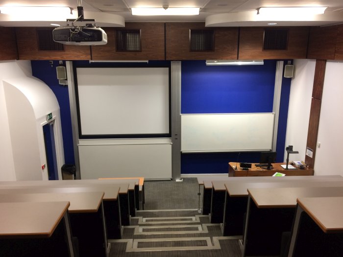Raked lecture theatre with fixed seating, projector, screens, whiteboards, and visualiser