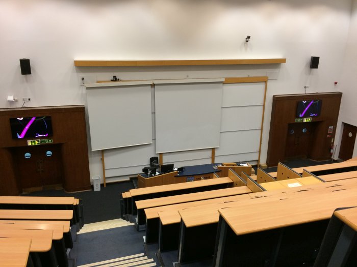 Raked lecture theatre with fixed seating, screens, whiteboards, and video monitors
