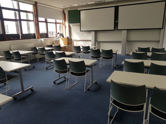 Flat floored teaching room with rows of tablet chairs, screens, chalkboards, and visualiser