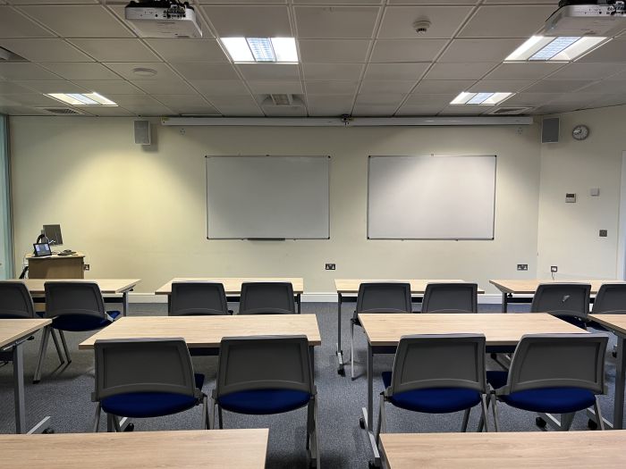 Flat floored teaching room with rows of tables and chairs, whiteboards, PC, and projector.