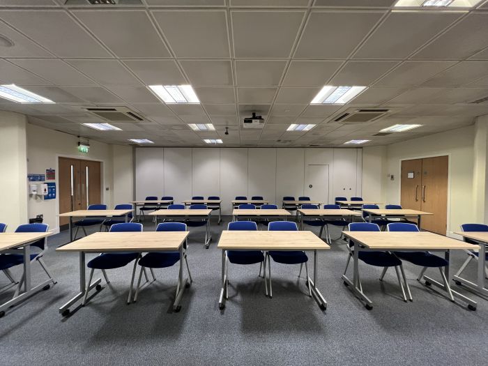Flat floored teaching room with groups of tables and chairs, projector, screen, and whiteboards
