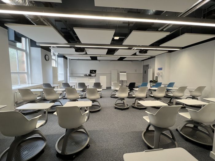 Flat floored teaching room with tablet chairs, moveable glassboard, moveable whiteboards PC, lectern, and projectors.
