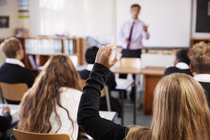 View from behind of classroom with a female pupil holding hand up to ask a question, 700px