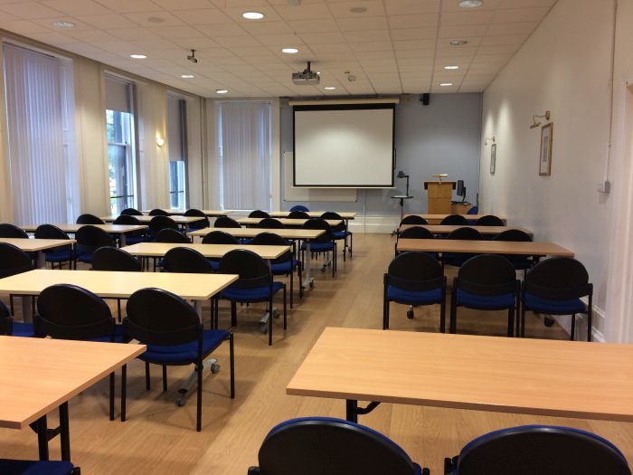 Flat floored teaching room with rows of tables and chairs, projector, screen, and whiteboard