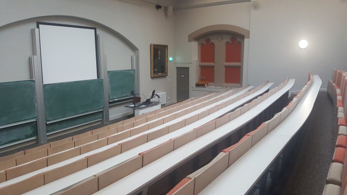 Raked lecture theatre with fixed seating, screen, chalkboards, and PC