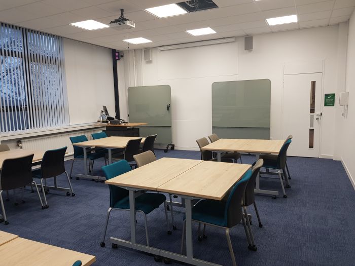 Flat floored teaching room with tables and chairs, screen, PC, lectern, and moveable glassboards.
