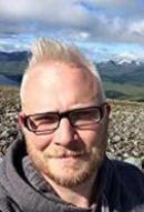 photo of Ben Colburn: headshot of an older balding white man with glasses and short facial hair, in a hoody. Wild scottish scsnery and overcast sky in the background