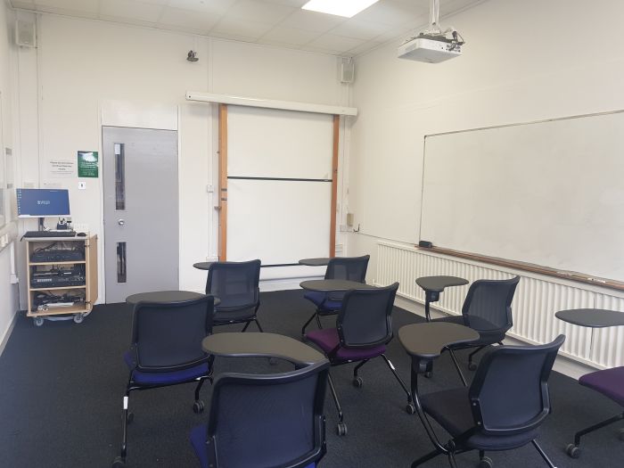 Flat floored teaching room with tablet chairs, whiteboard, PC, and projector.