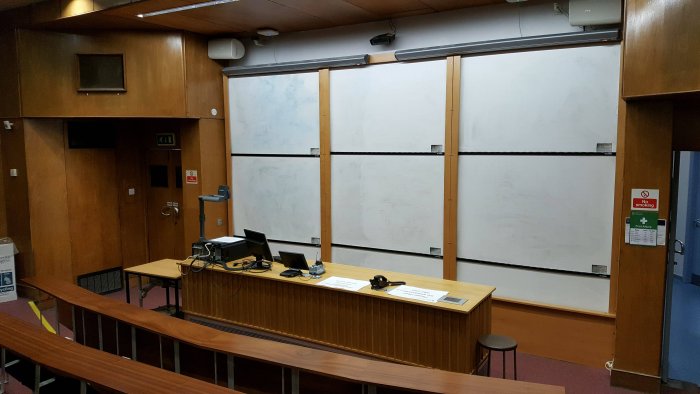 Raked lecture theatre with fixed seating, whiteboards, screens, visualiser, and PC