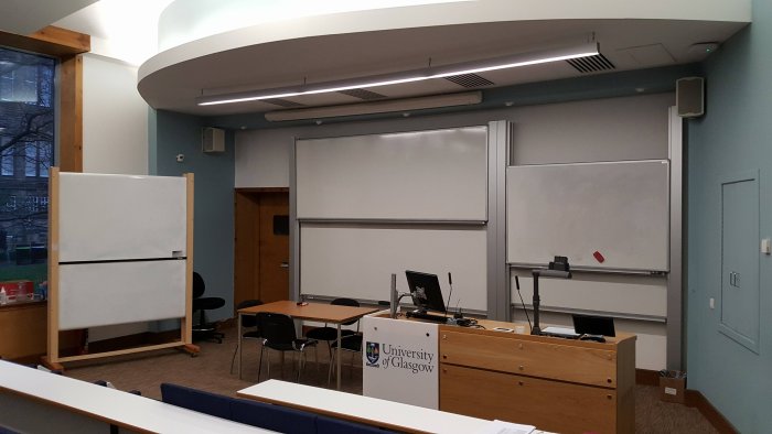 Raked lecture theatre with fixed seating, whiteboards, screens, visualiser, and PC