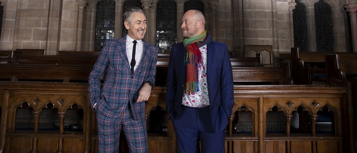A photo of the actor Alan Cumming and theatre director John Tiffany leaning on a wooden bench in the University's Bute Hall