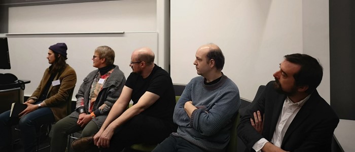 photo of a line of white middle-aged people (four men and one woman) sitting on chairs, all looking to the left