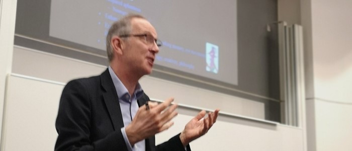 photo of an older white man with grey receding hairline, glasses, blue shirt and suit jacket gesticulating while speaking in front of a projection screen