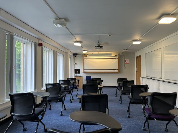 Flat floored teaching room with tables and chairs in round table set-up, whiteboard, screen, projector, and PC