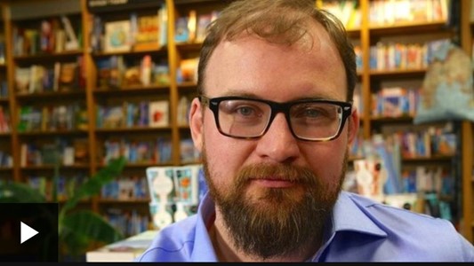 close-up photo of a white man in his 30s with red hair and mid-length beard and black glasses, in a library