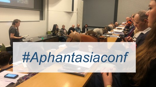 photograph of a conference q&A panel headed by a white woman, standing up in front of a projector and desk, with participants at the panel as well as audience (white people of mixed genders, all over 30) visible in foreground. Overlaid in a white box is text hashtag aphantasiaconf