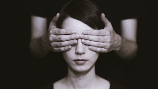 black and white close-up photo of a white woman with black hair with a pair of white hands over her eyes, belonging to a person behind her in a dark t-shirt