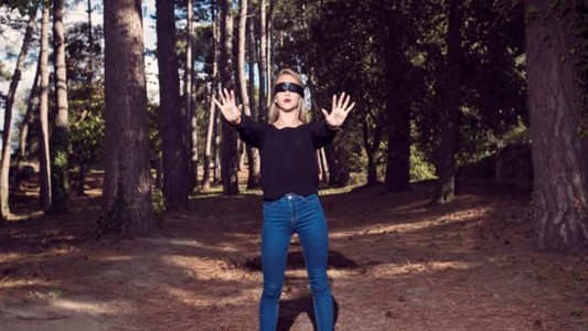 a photograph of a white woman dressed in black top and jeans skirt, a black scarf tied around her eyes, stumbing with her hands extended towards the viewer in a well-lit forest