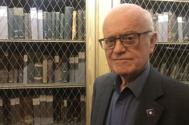A portrait photo of Professor Dugald Cameron in front of shelves of books kept behind glass doors