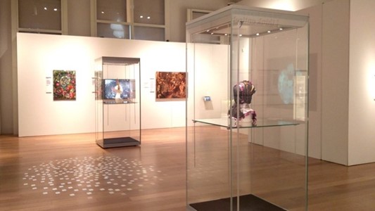 photo of the inside of a gallery with well-lit white walls on which framed artworks hang, as well as two plexiglass cases with two busts exhibiting artwork by Claire Strickland.