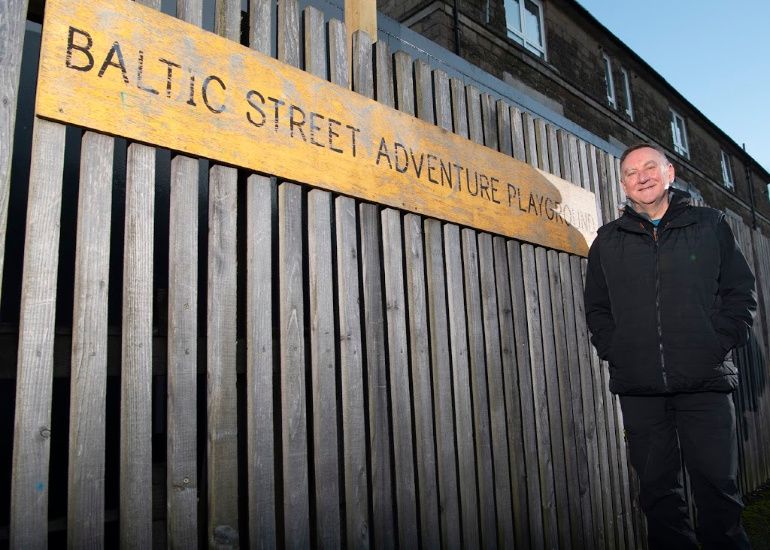 Photo of Alistair McCall, one of the play workers from Baltic Street Adventure Playground, next to the Baltic Street Adventure Playground sign