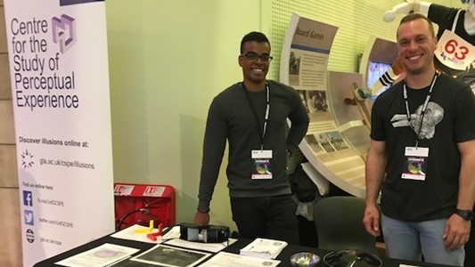 photo of two young men (a black man in dark clothes and a white man in a black jumper with a rabbit on it) behind a table full of cards and documents. On their right (left of the picture) a banner much taller than the men with a purple logo and purple text: 'Centre for the study of perceptual experience'