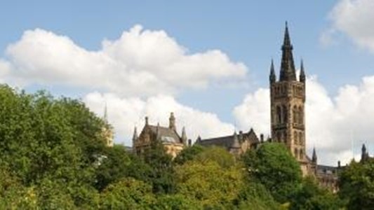 photo of outdoors: a panoramic view of a grey gothic tower (the university of Glasgow) with lush greenery in the foreground and blue sky with white fluffy clouds in the background